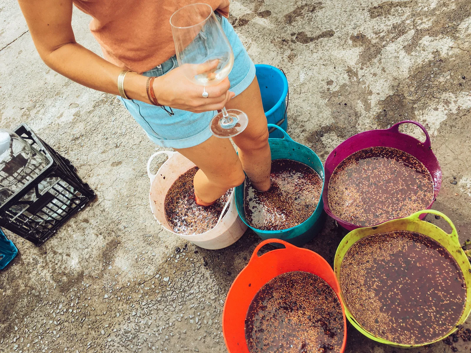 Crushed grapes in coloured tubs with somebody standing in one of them, a glass of wine in hand
