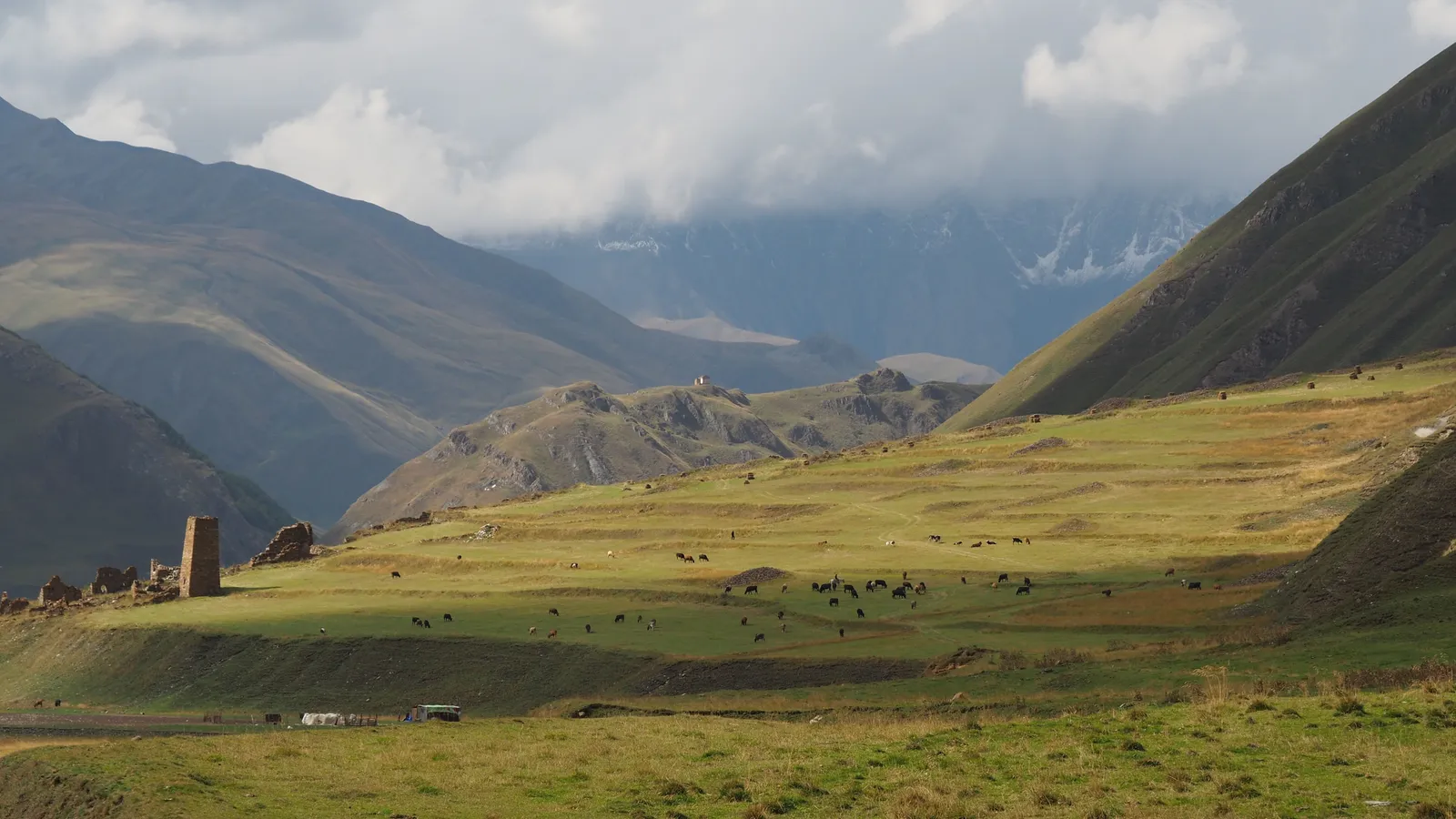The broad pasture floor of the Truso valley with an abandoned stone tower on the slope and snow peaks at its head