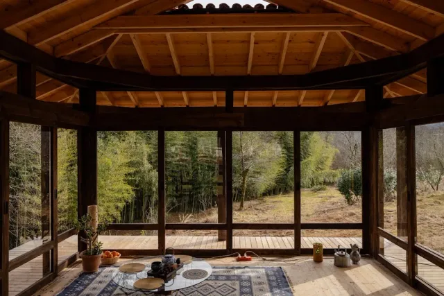 A wooden pavilion with exposed beams and glass walls, rugs and a low table facing a bamboo grove