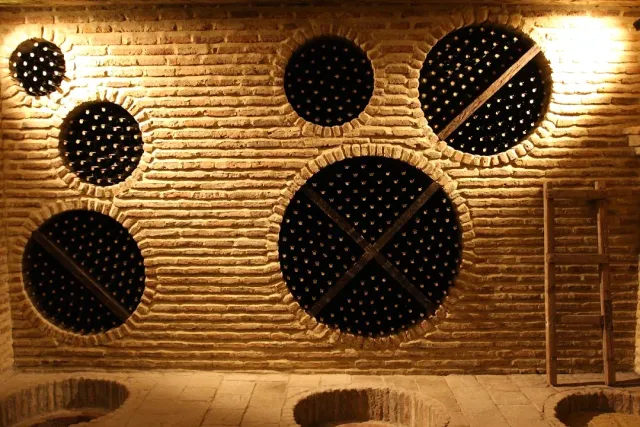 A stone wall with round recesses full of wine bottles in warm light and jar openings in the floor