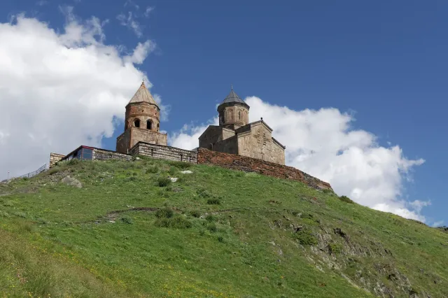 A stone church with a conical dome and a separate bell tower on a green ridge under clouds