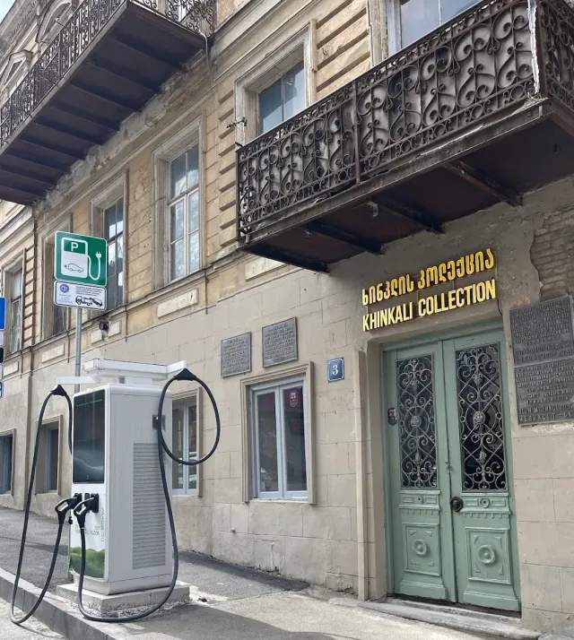 A stone building with ornate iron balconies, a green wooden door and a charging point on the pavement