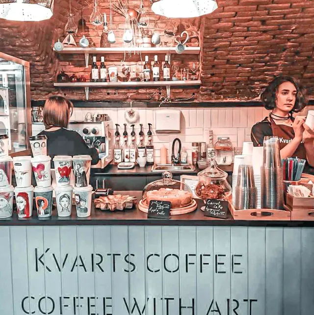A counter with two baristas, illustrated paper cups, cakes under a glass dome and a brick wall