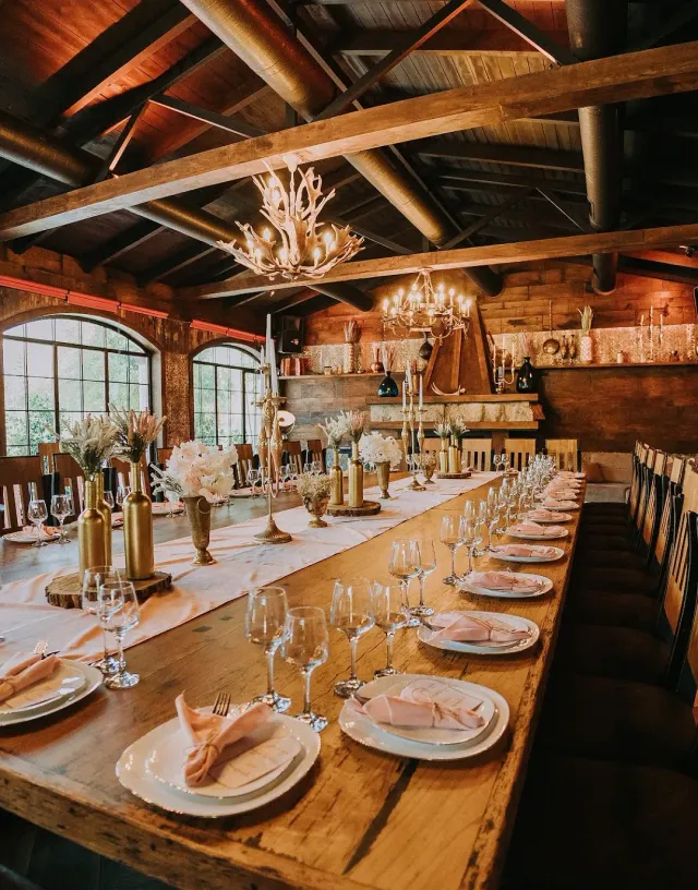 A long table set with pink napkins under wooden beams, with an antler chandelier and arched windows