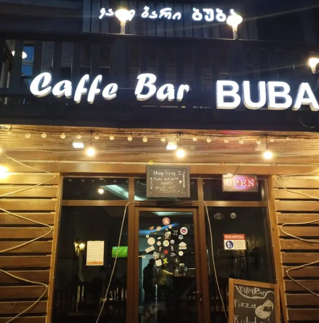 A wooden facade at night with a lit sign, a string of bulbs and a glass entrance covered in stickers