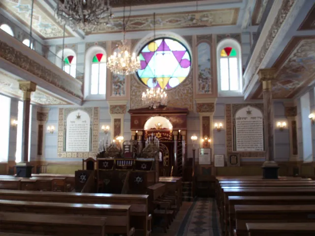 A prayer hall with wooden benches, a central chandelier and a Star of David stained glass window