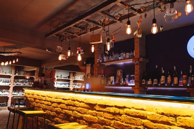A stone counter lit by a yellow light strip, bulbs hanging above it and bottle shelves behind
