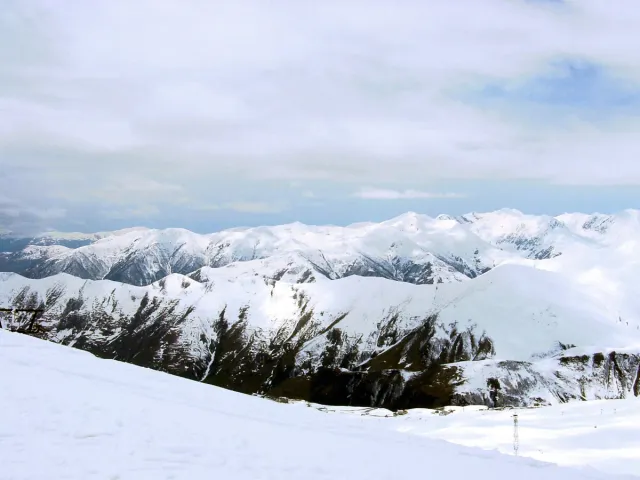 A broad snow slope facing snow covered mountain ridges with scattered fir trees