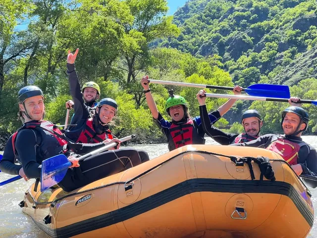 A group with helmets and paddles cheering in a rubber raft on a river between forested mountains