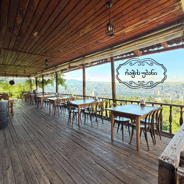 A covered wooden terrace with tables and rounded chairs, and behind a view of a city and mountains