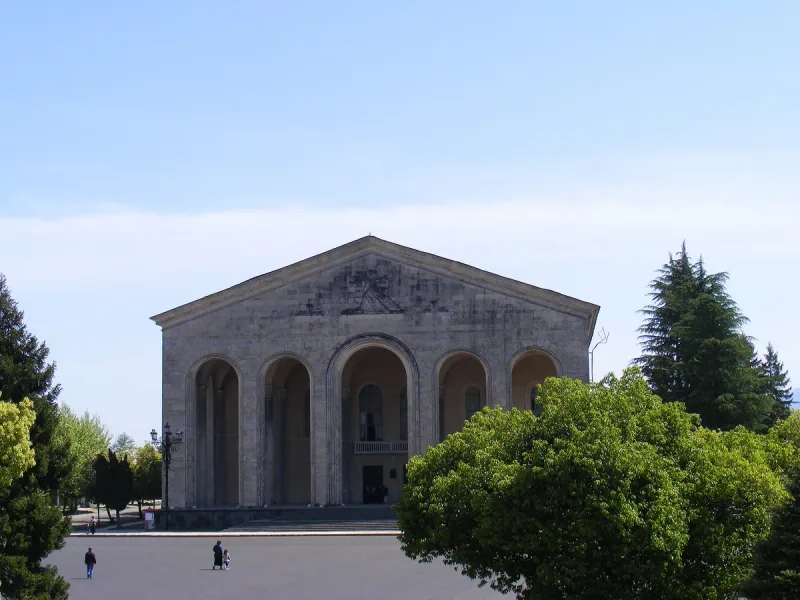 The stone facade of Ozurgeti Drama Theater with its row of arches and columns, framed by trees under a blue sky