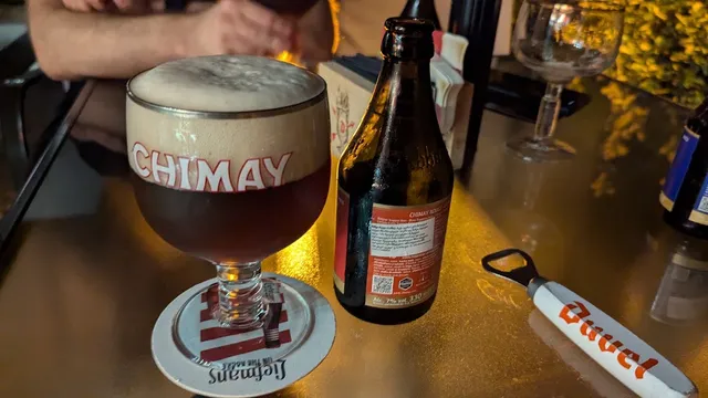 A glass of dark beer with foam and a bottle beside it on a table in warm light