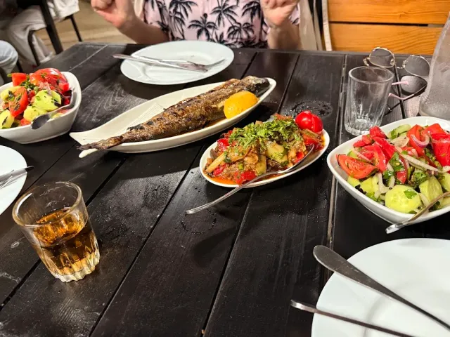 A whole grilled fish with a lemon wedge on a dark table, beside bowls of vegetable salad