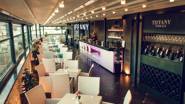 A long glazed terrace with white wicker armchairs, a lit counter and a wall of wine bottles