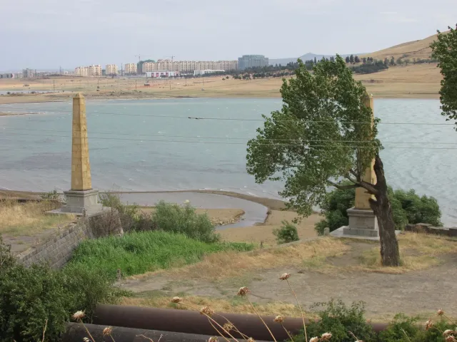 The shore of a wide reservoir with trees and a stone monument on the promenade