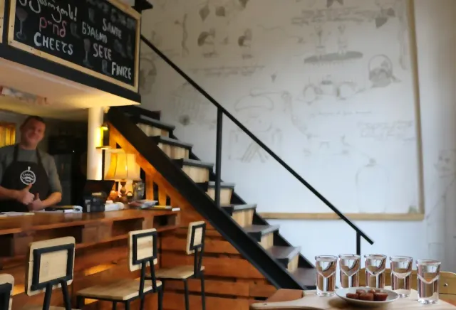 A bartender behind a wooden counter with stairs beside it, and five shot glasses on the table in front