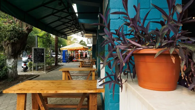 Light wooden tables along a turquoise wall and a large planter facing a shaded sidewalk