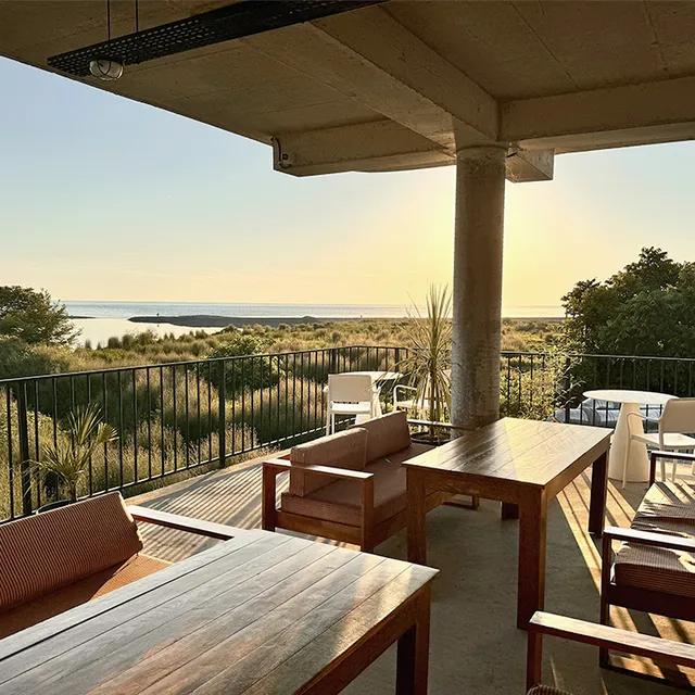 A covered terrace with sofas and wooden tables, facing coastal vegetation and the sea at sunset