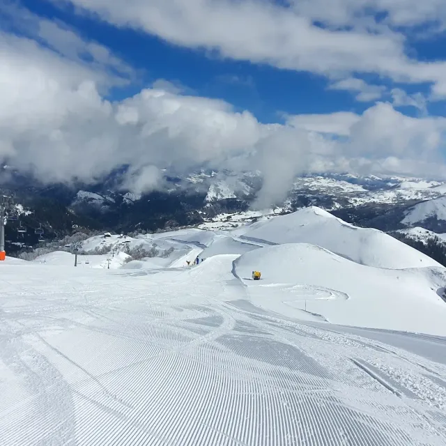 A groomed ski slope in the snow with snowy mountains and low clouds on the horizon
