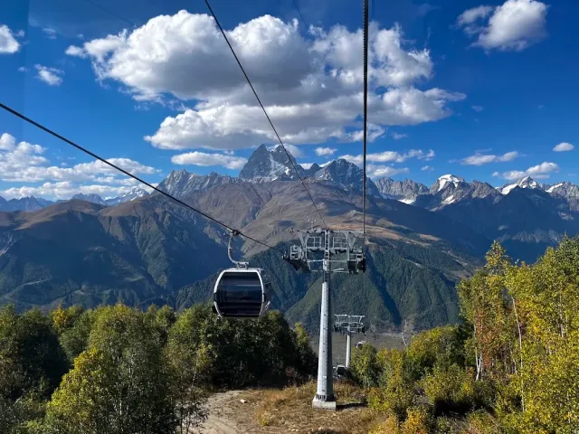 A gondola cabin on the cable facing snow capped peaks, blue sky and trees in autumn colors