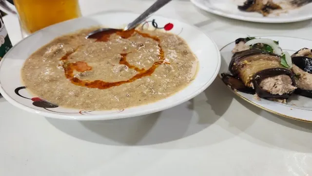 A bowl of thick soup with a line of red oil, beside a plate of eggplant rolls and a glass of beer