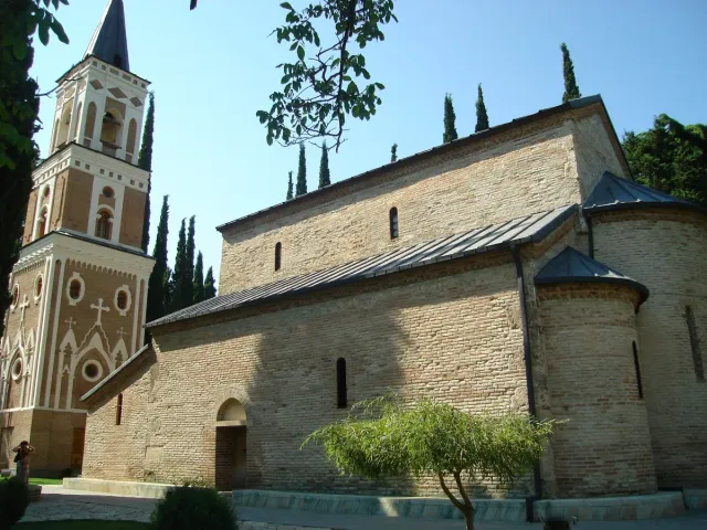 A stone basilica beside a brick patterned bell tower with a row of cypresses