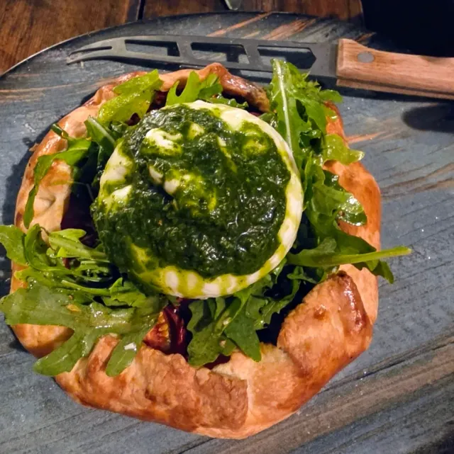 An open pastry with rocket leaves and a round cheese covered in green sauce, on a dark board