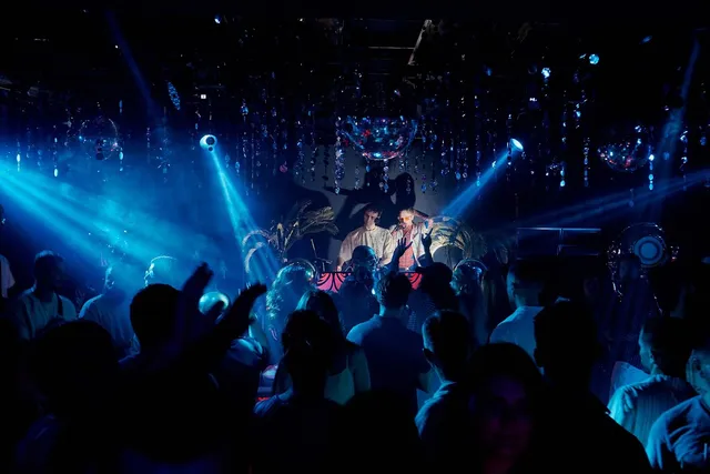 A crowded dance floor facing a DJ booth in blue lights and mirror balls