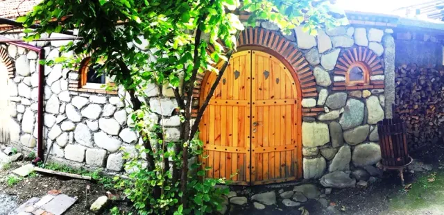 An arched wooden gate in a river stone wall, with a climbing vine and stacked firewood at the side