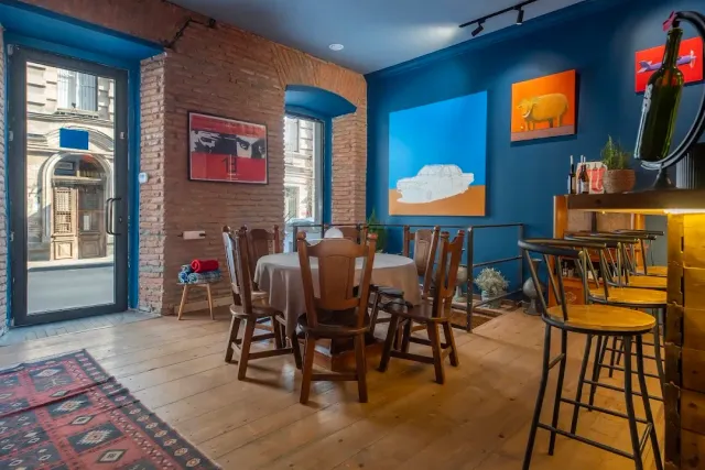 A room with blue walls and exposed brick, a round table, colourful paintings and tall stools
