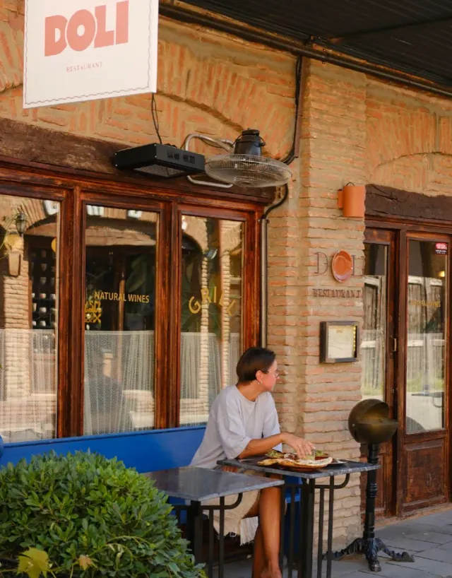 A person sitting at a small marble table by a brick facade with wooden windows and a hanging sign