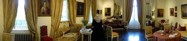 A historic drawing room with a grand piano, gold curtains, portraits and a period dress