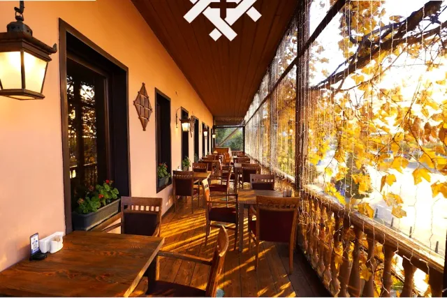 A covered wooden balcony with tables and chairs, lanterns and yellow foliage beyond a net