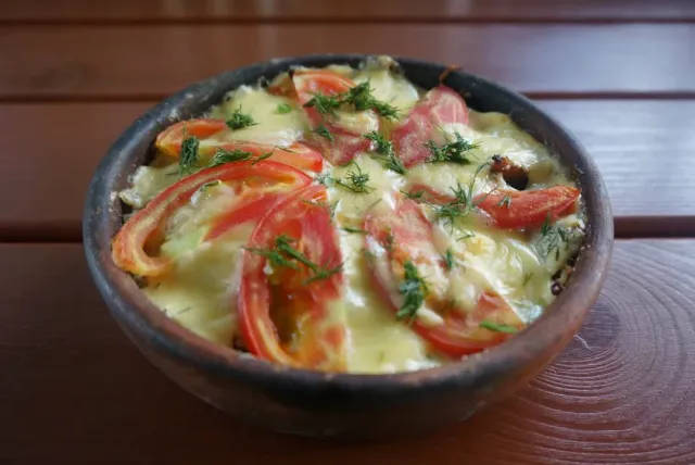 A clay bowl with a dish baked in melted cheese, tomato slices and dill on a wooden table