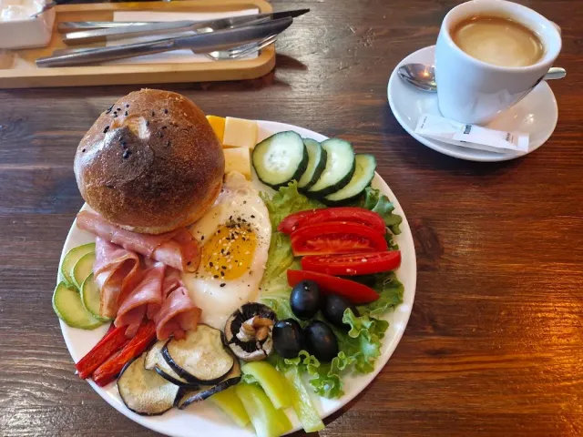 A breakfast plate with a bun, a fried egg, slices of meat and cut vegetables beside a coffee cup