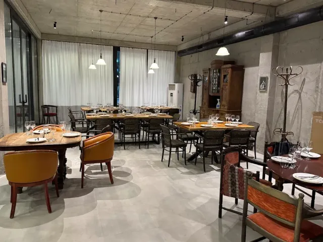 A room with a concrete ceiling, light curtains, set tables, mustard armchairs and a wooden cabinet