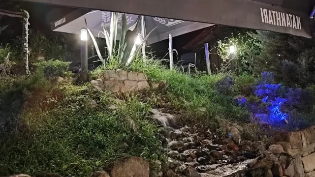 A rock garden lit at night with a small stream and a canopy over tables in the background