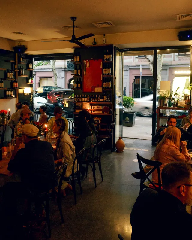 A space full of diners in the evening with wine bottle shelves and doors open to the street