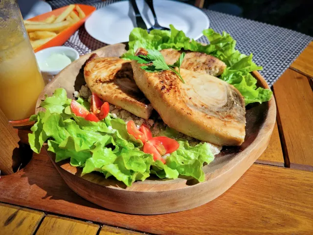 A wooden bowl with slices of grilled fish on lettuce and tomatoes, beside fried potatoes
