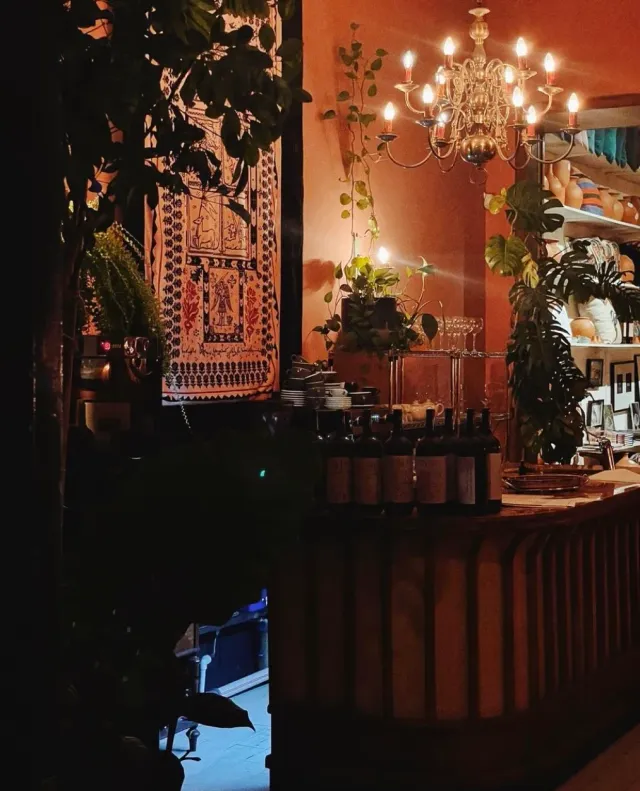 A dim warm space with a candle chandelier, a wall rug, plants and a row of bottles on a wooden counter
