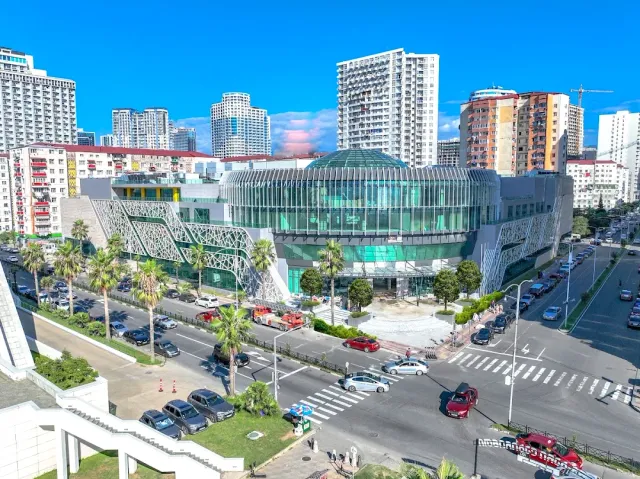An elevated view of a glass mall building with a green lattice facade, palms and a busy road