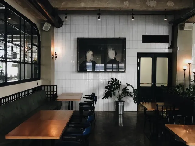 A space with a white tiled wall, a large framed photograph, wooden tables and black chairs