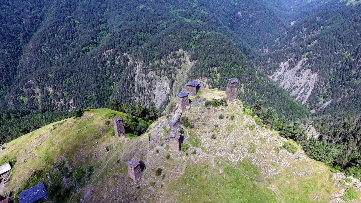 Stone defensive towers on a grassy ridge above a forested valley, seen from above