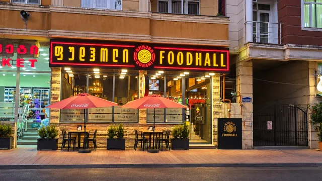 A facade lit in the evening with a red neon sign, red umbrellas and tables on the sidewalk