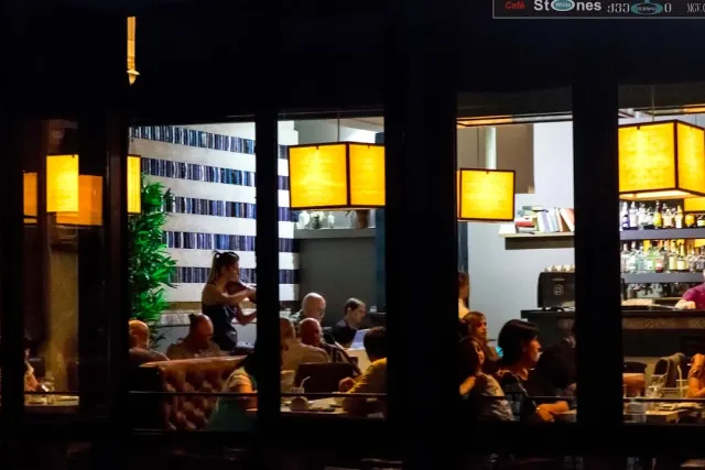 A view from the street into a space lit by yellow ceiling lamps, with diners at the tables and a bar