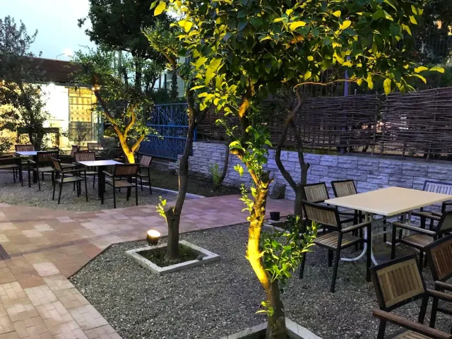 A paved courtyard in the evening with tables, trees lit from below and a wicker fence