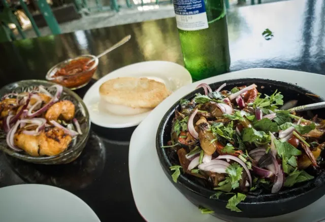 A stone bowl with pieces of meat, coriander and red onion, beside sauce, bread and a green bottle