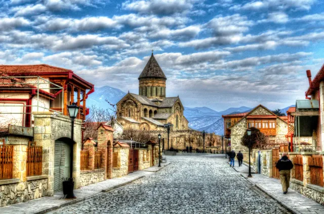A paved street of low houses with wooden balconies and a church dome at its end