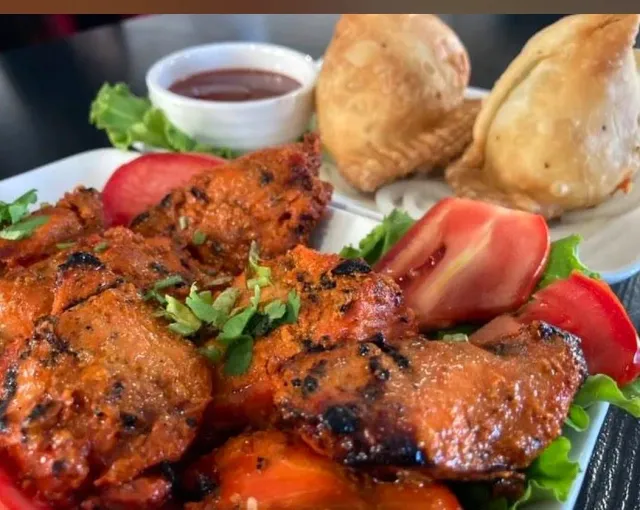 A plate of red chicken pieces with lettuce leaves, tomato slices, triangular pastries and sauce