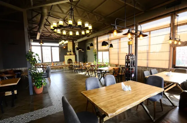 A restaurant hall with wooden tables, candle chandeliers and a wine rack, in daylight through the blinds